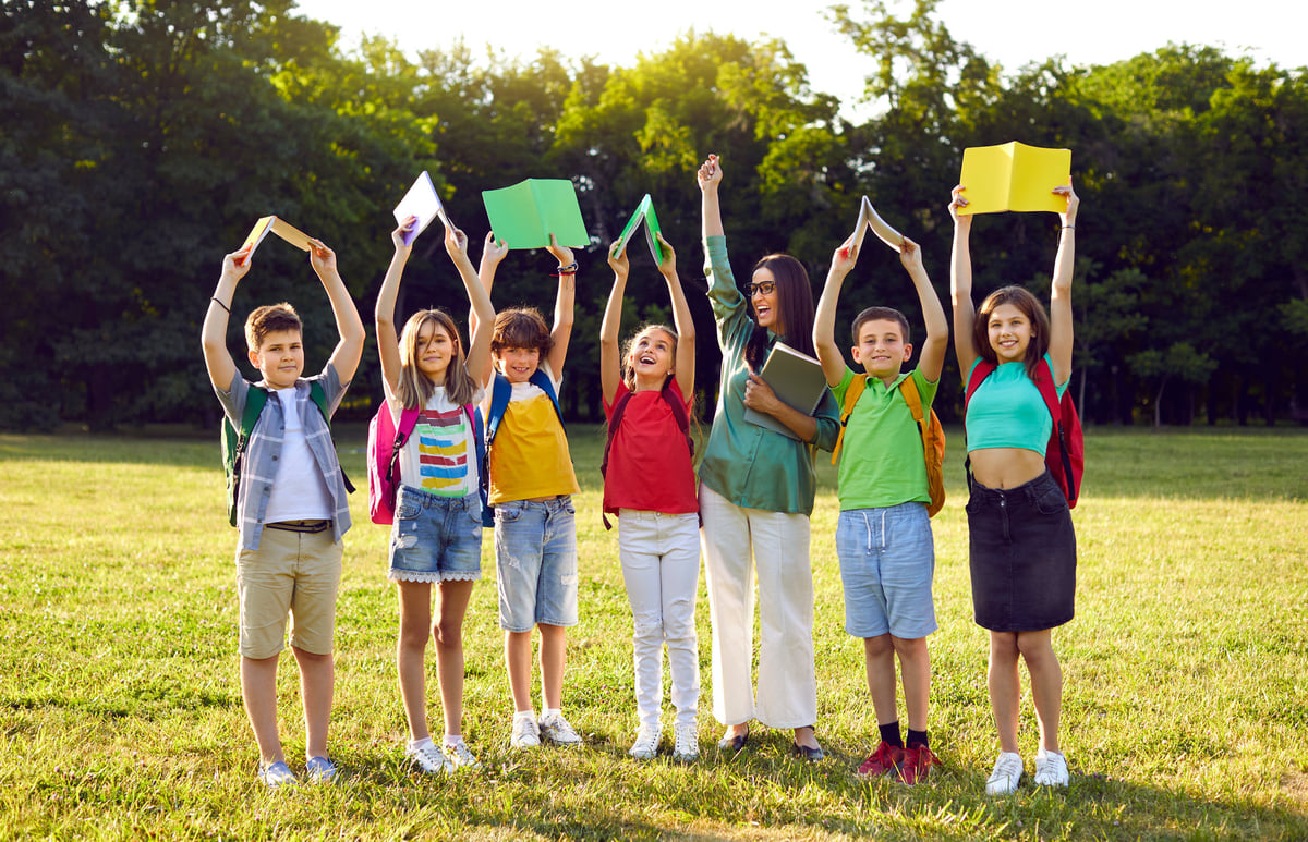 Female teacher and students having fun interesting class outside classroom. Portrait of happy, excited school teacher together with children standing in row in green field or schoolyard
